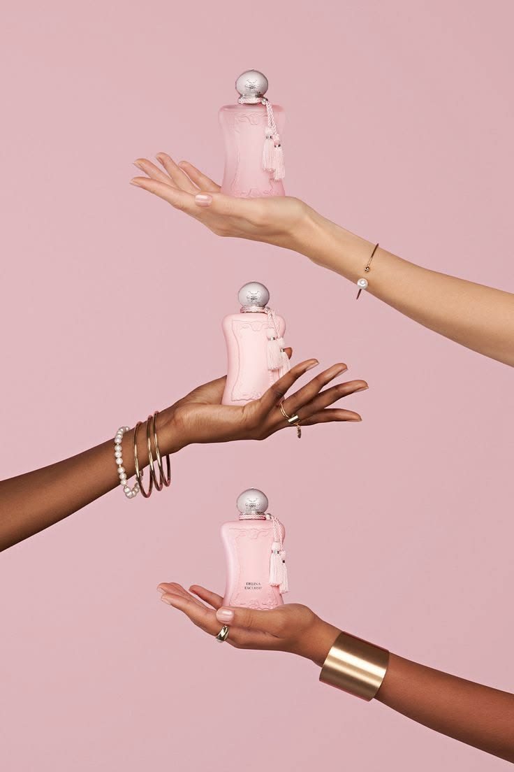 Three hands each holding a pink Parfums de Marly Delina bottle against a pink background