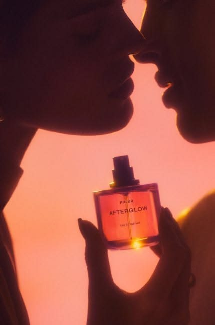 Two faces leaning close together in warm orange light with a bottle of Phlur Afterglow held between them